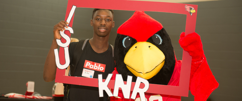 A Student, holding a red frame decorated with the letters I S U. K N R, posing with Reggie Redbird.